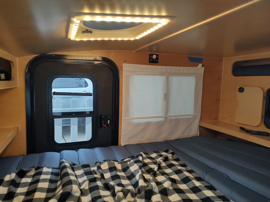 Compact RV rental interior featuring a comfortable bed with plaid blanket, a door, window with shade, and overhead LED lighting.