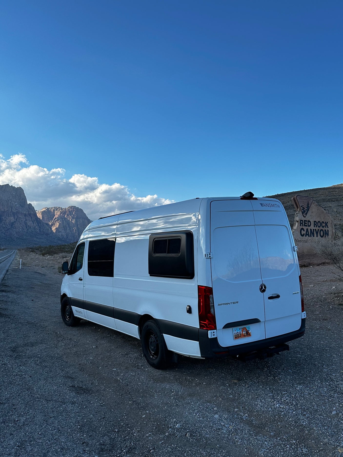 White Sprinter RV rental van parked on gravel at Red Rock Canyon, Nevada, with scenic mountains under a blue sky.