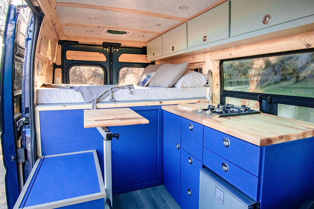 Vibrant blue interior of this RV rental camper van boasts a raised bed, full kitchen, and scenic window view.