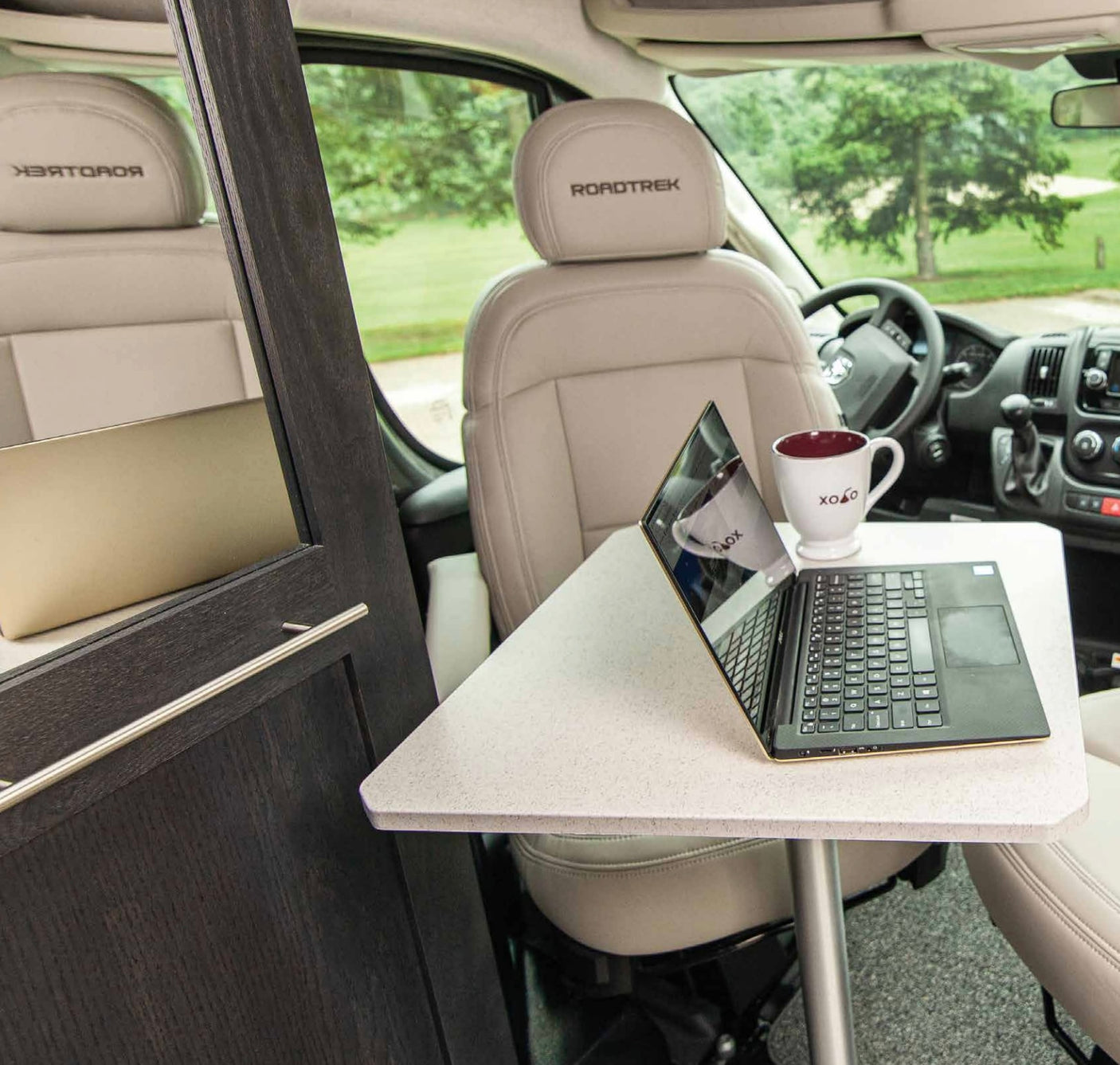 Work on the road! This RV rental offers a cozy mobile office with "ROADTREK" seats, a laptop, and a mug on a table.