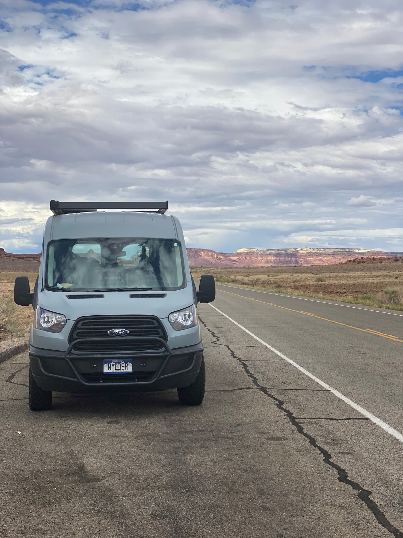 An RV rental, a grey Ford Transit camper van, is parked on a scenic desert road with distant red cliffs and a cloudy sky.