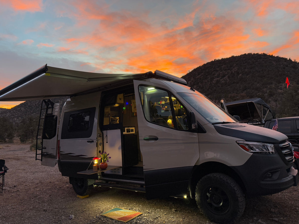 Gorgeous sunset over an RV rental camper van campsite. Awning extended, lights on, revealing a welcoming outdoor setup.