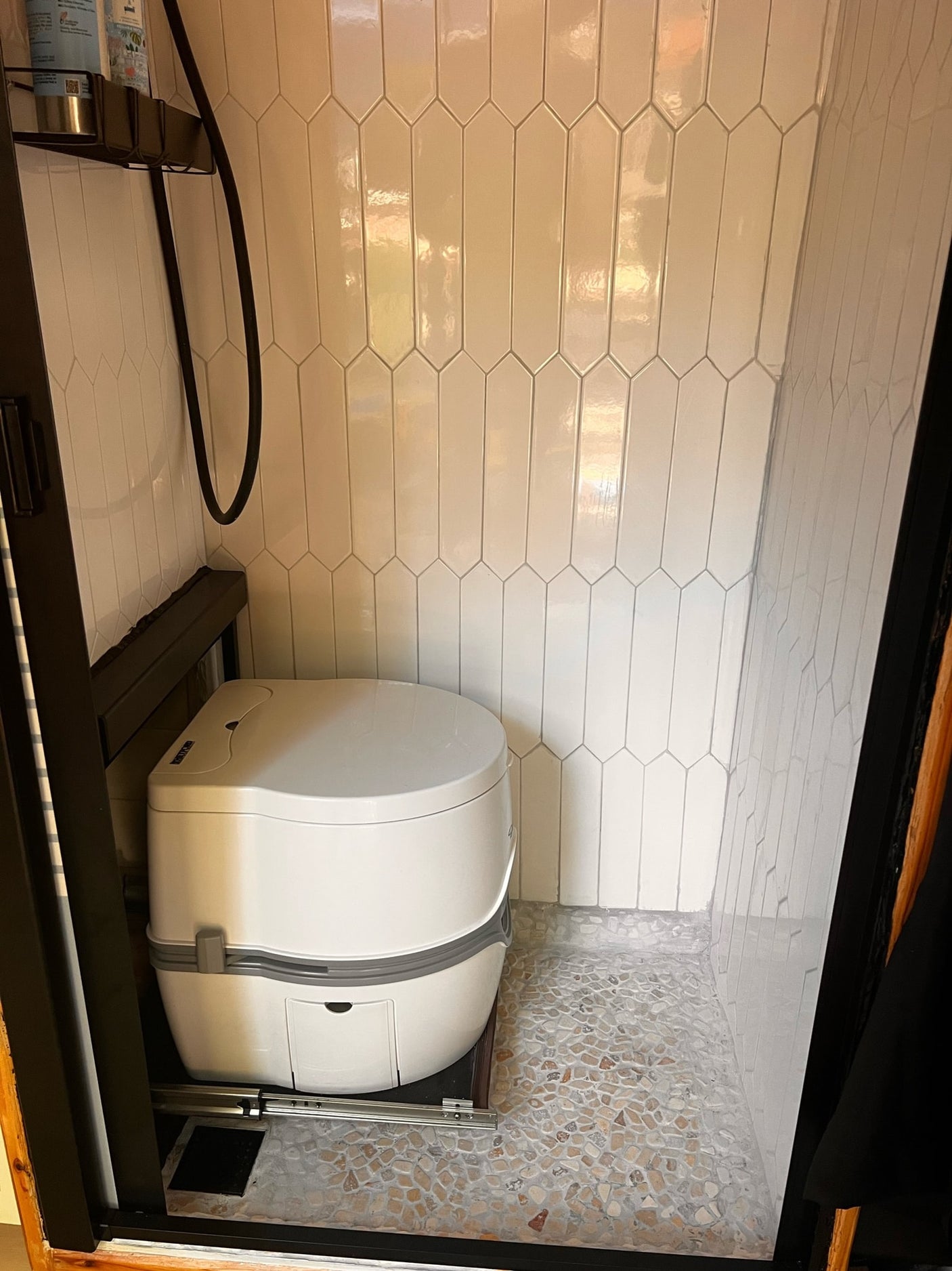 Modern RV rental bathroom: pull-out composting toilet, white tiled walls, and pebble floor shower.