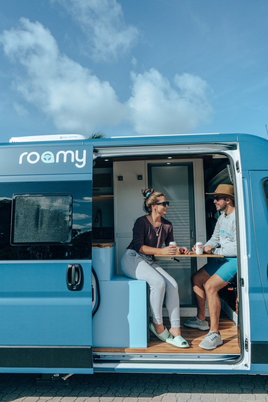 A couple relaxes with drinks inside their blue Roamy RV rental, showcasing the comfortable campervan interior.