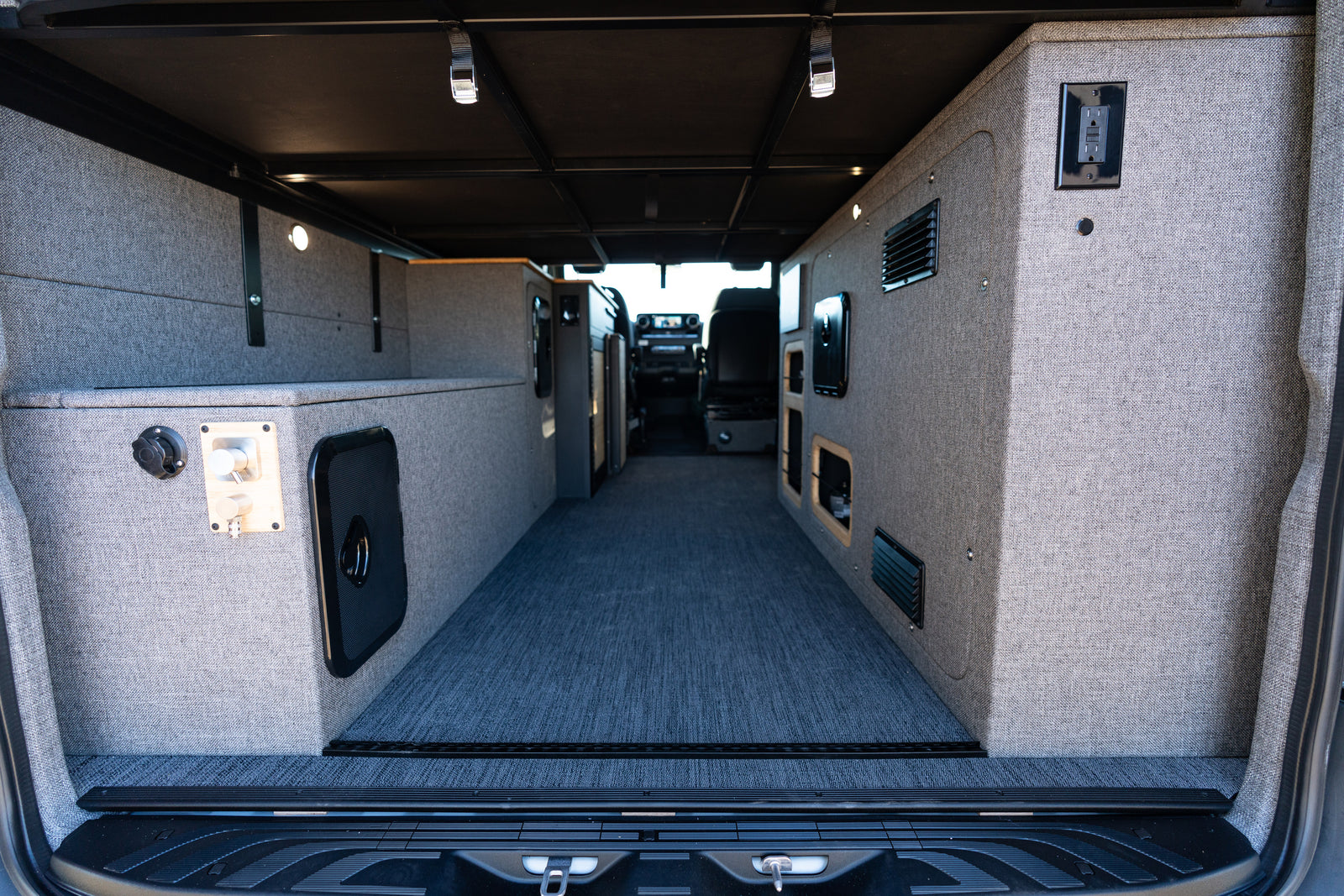 Custom RV rental interior features gray upholstered walls, integrated storage, carpet, and lighting, viewed from rear.