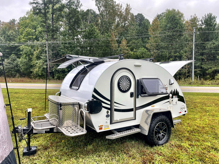parked on grass with an open window and awning on a cloudy day.|A modern white & black teardrop RV rental