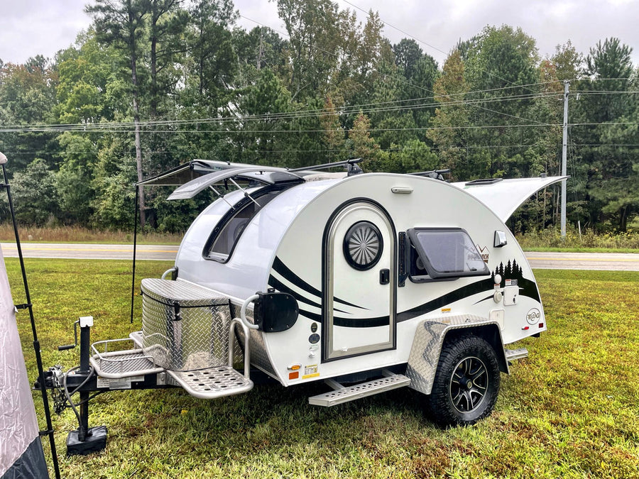 parked on grass with an open window and awning on a cloudy day.|A modern white & black teardrop RV rental
