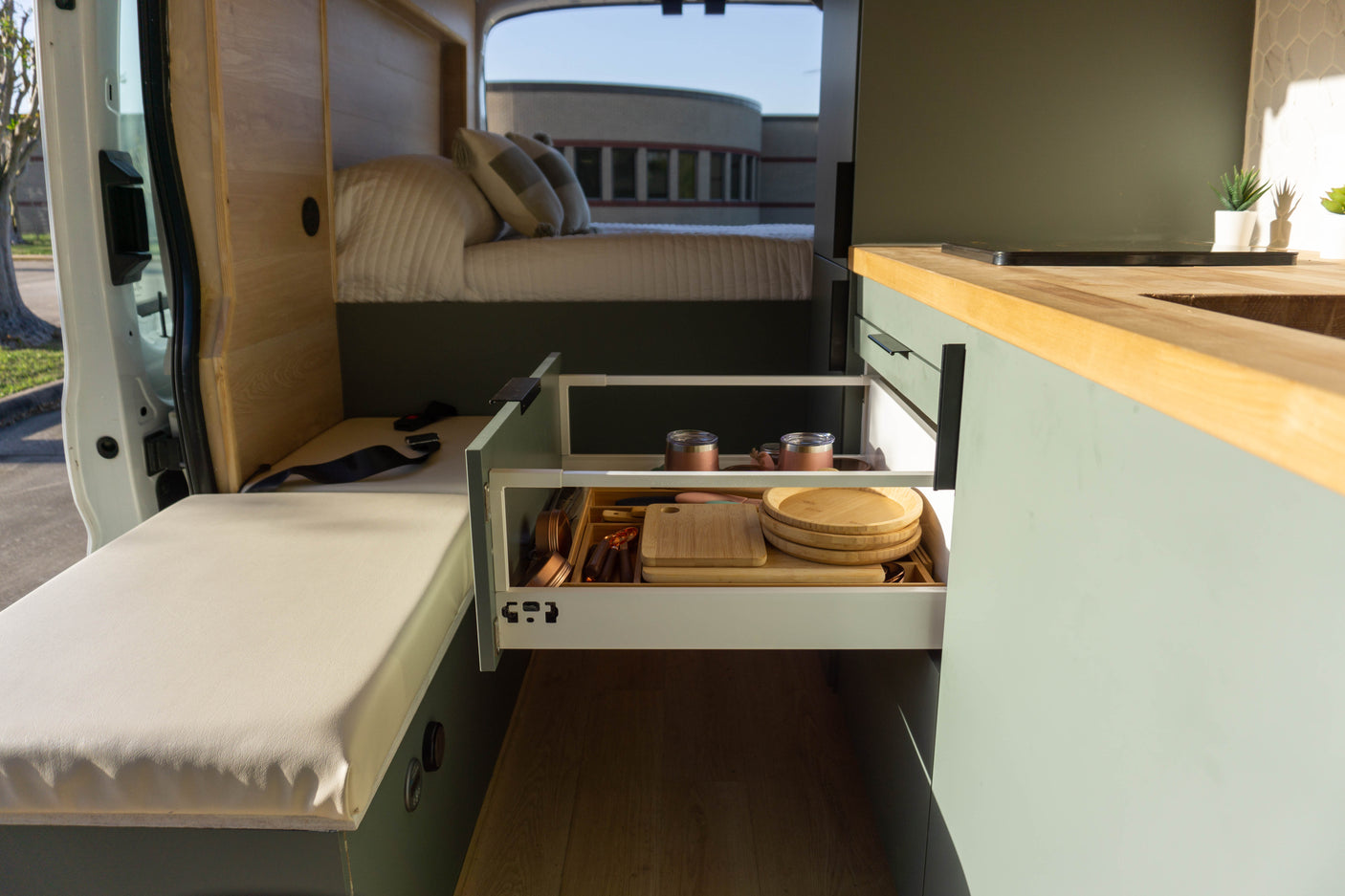 Modern RV rental interior: an open kitchen drawer displays dishes, utensils, and cutting boards; a cozy bed is visible.