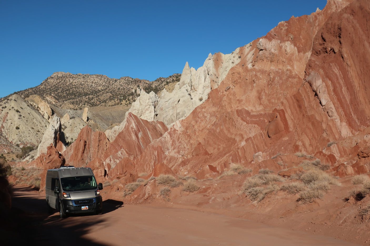A grey RV rental van travels on a red dirt road, surrounded by stunning, colorful rock formations and mountains under a blue sky.