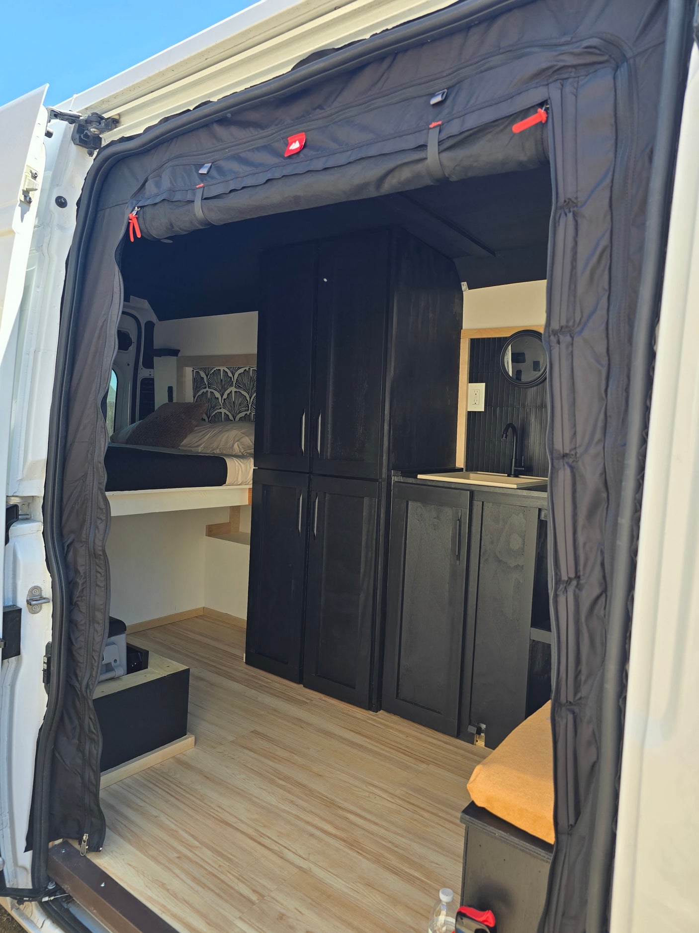 Inside this RV rental: light wood flooring, black cabinets, a sink, comfy bed, and door privacy curtains.
