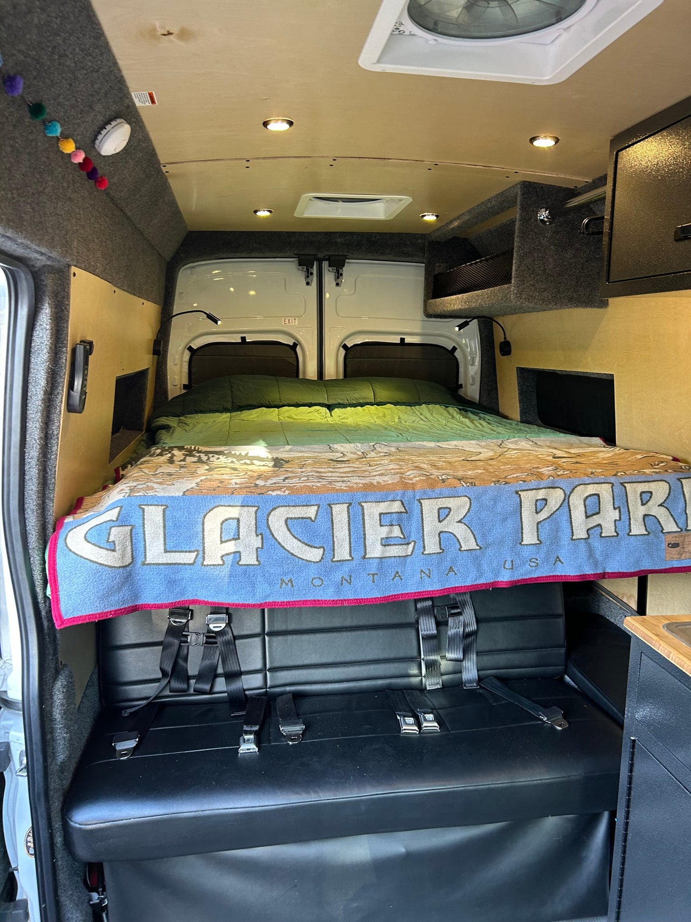 View inside an RV rental showing a bed with a "Glacier Park" blanket, positioned above a bench seat with belts. — photo 7