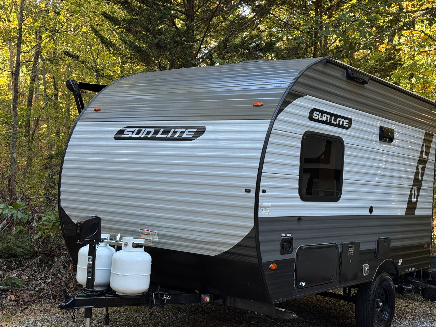 Compact Sun Lite travel trailer RV rental, featuring two propane tanks, parked in a lush, green wooded setting.