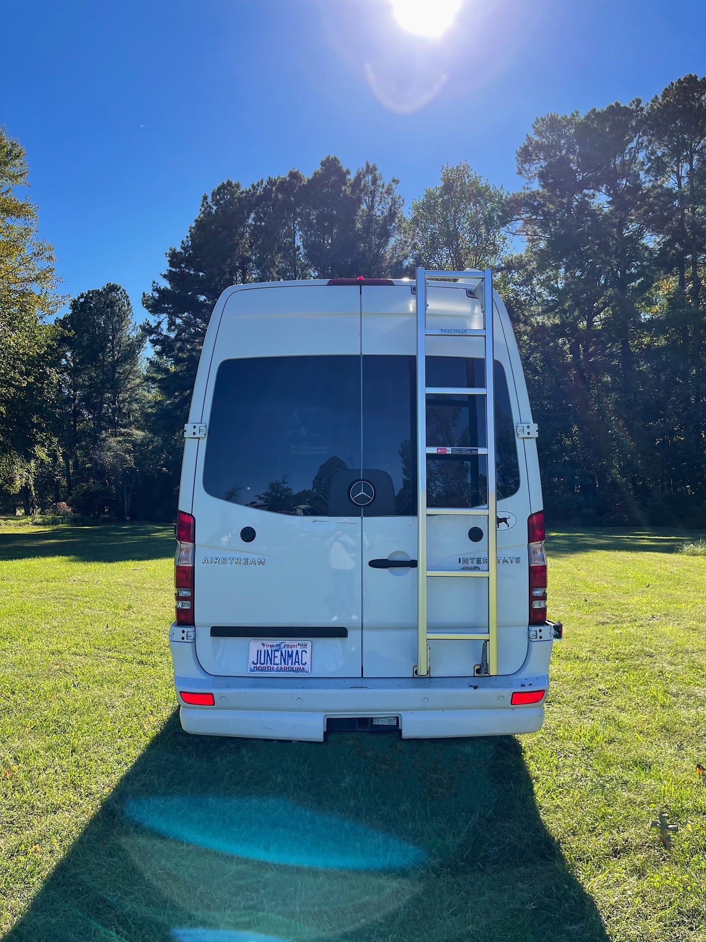 A white Airstream Interstate RV rental, seen from the rear with a ladder, sits on a sunny green lawn surrounded by trees.