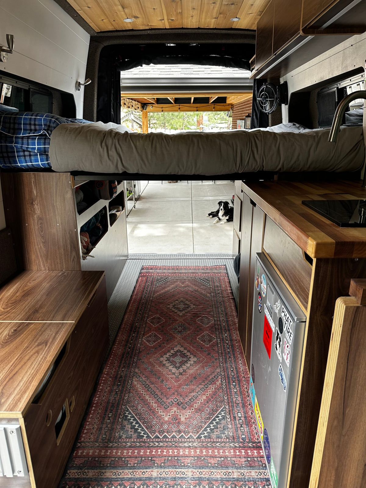 Cozy RV rental interior featuring a raised bed, kitchen, custom cabinetry, a patterned rug, and a dog relaxing outside. — photo 12