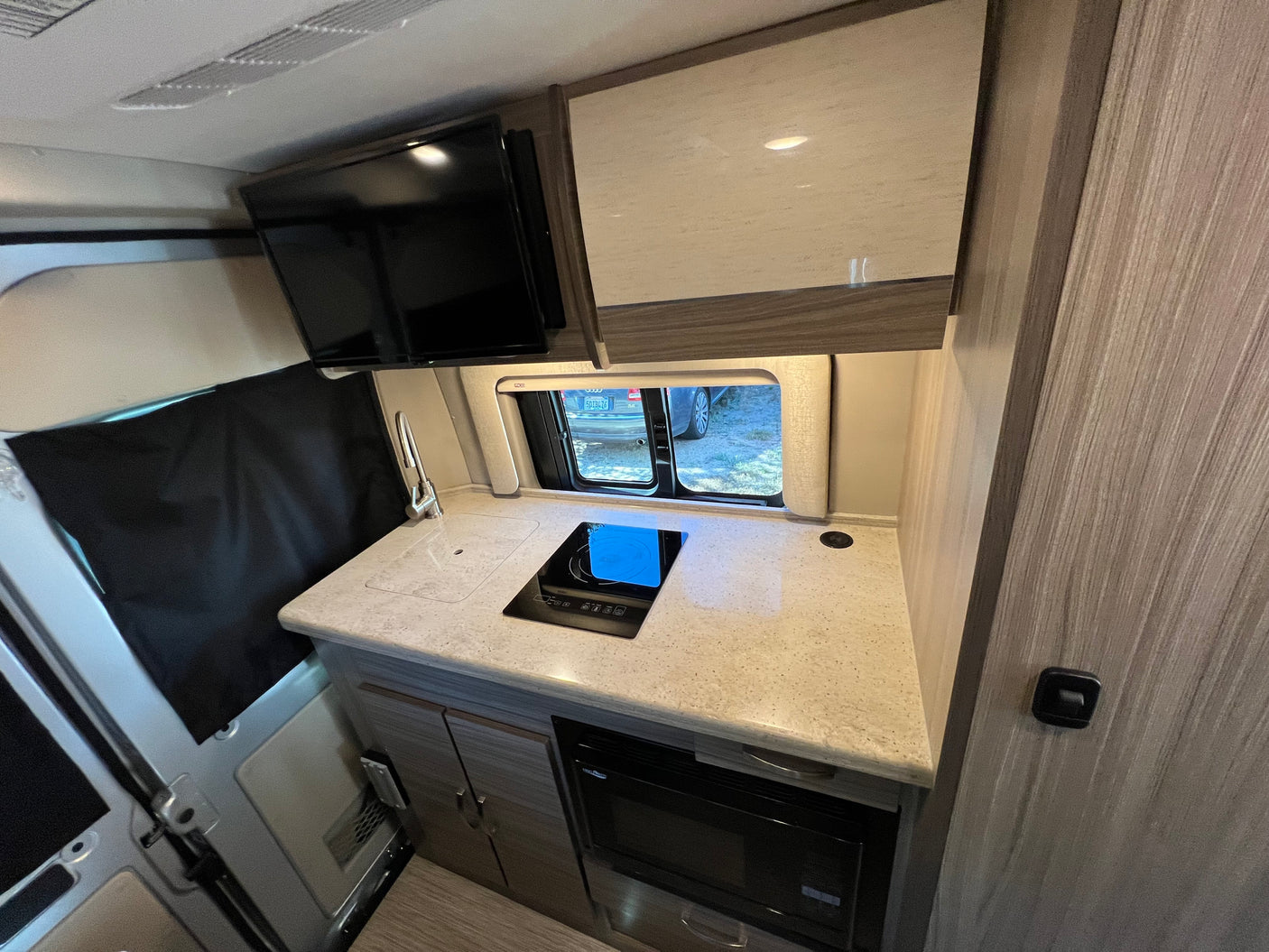 RV rental kitchen: counter, sink, induction cooktop, microwave, TV, and wood-grain cabinets.