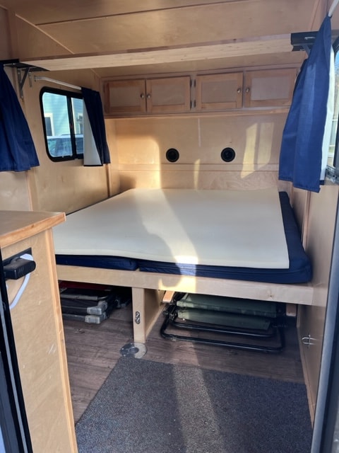 RV rental interior: view of a light wood cabin with a bed, storage cabinets, and stowed folding chairs.