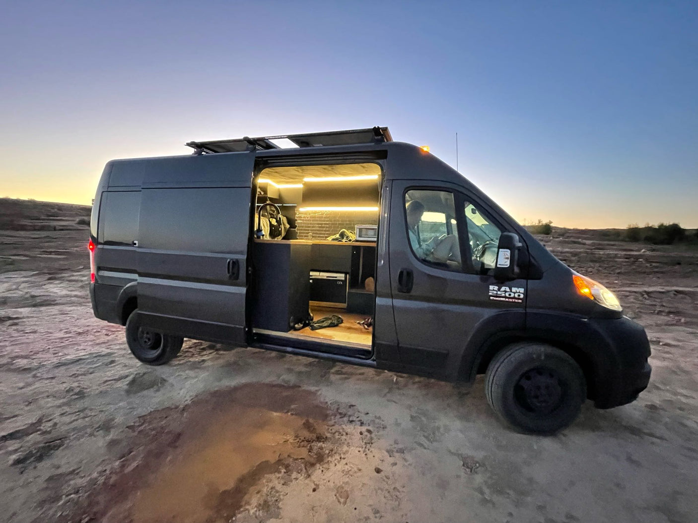 Experience off-grid van life! This custom-converted Ram ProMaster RV rental, with its cozy interior, is parked at dusk in a desert setting.