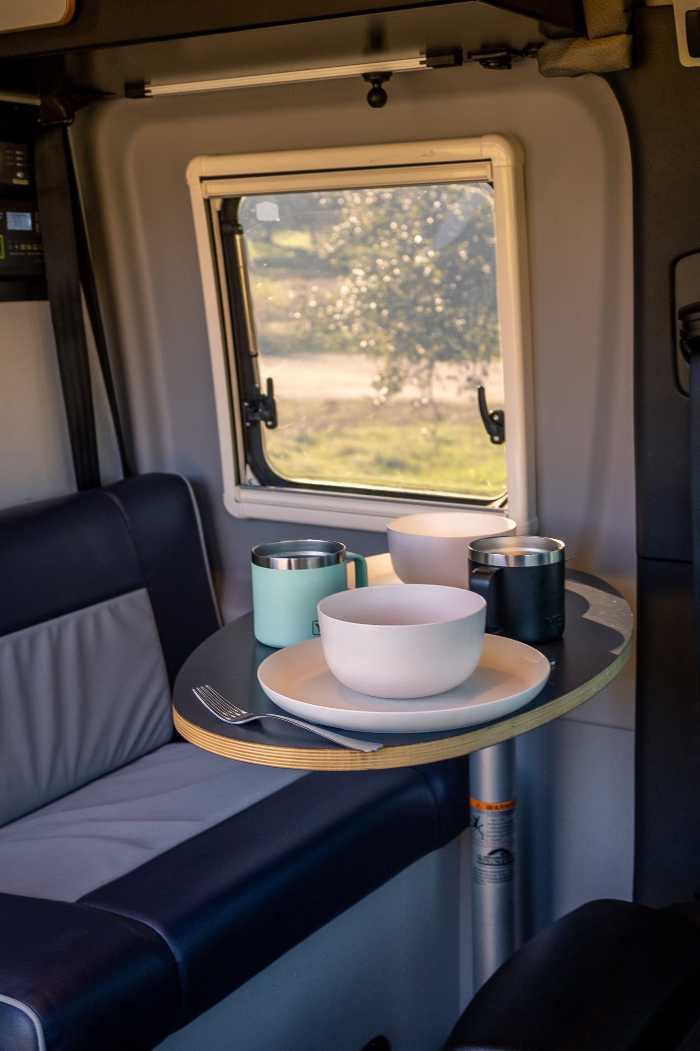 RV rental interior: A dining table holds plates, bowls, and mugs by a window view, perfect for camper van travels.