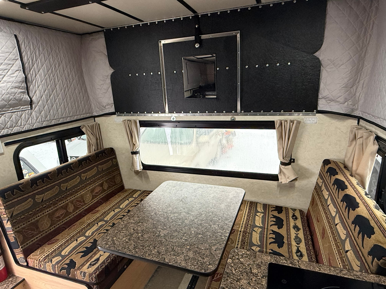 An rv rental camper interior showcasing a cozy dinette with bear and canoe patterned benches, a table, and large window with curtains.