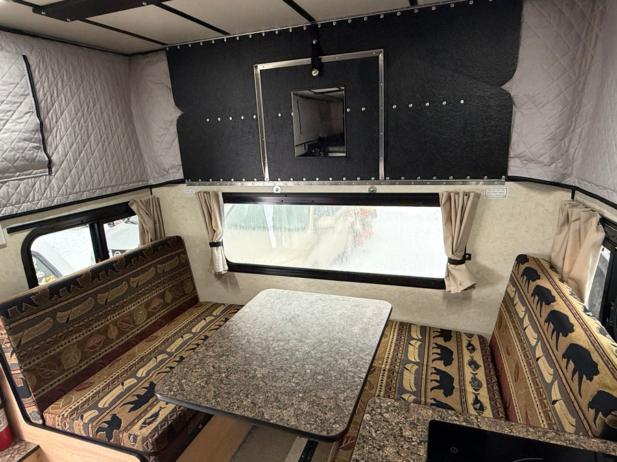 An rv rental camper interior showcasing a cozy dinette with bear and canoe patterned benches, a table, and large window with curtains.