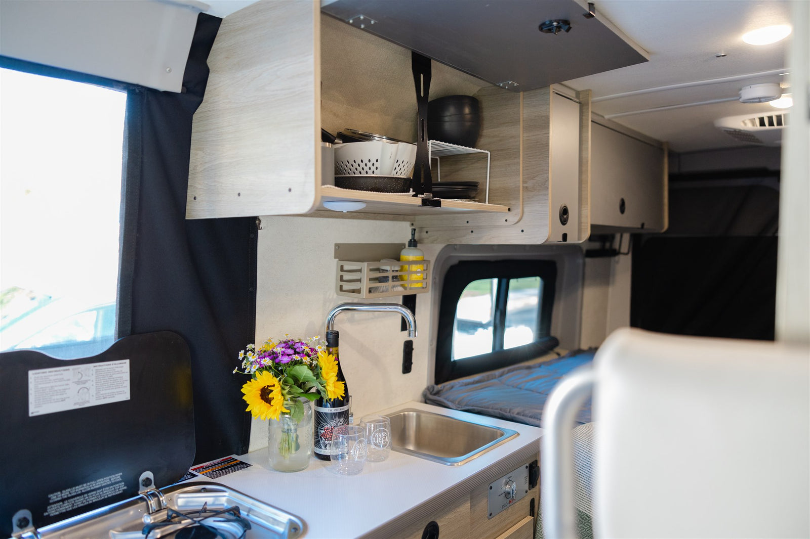 Well-equipped rv rental interior: compact kitchen galley with sink, stove, overhead storage, and flowers on the counter.