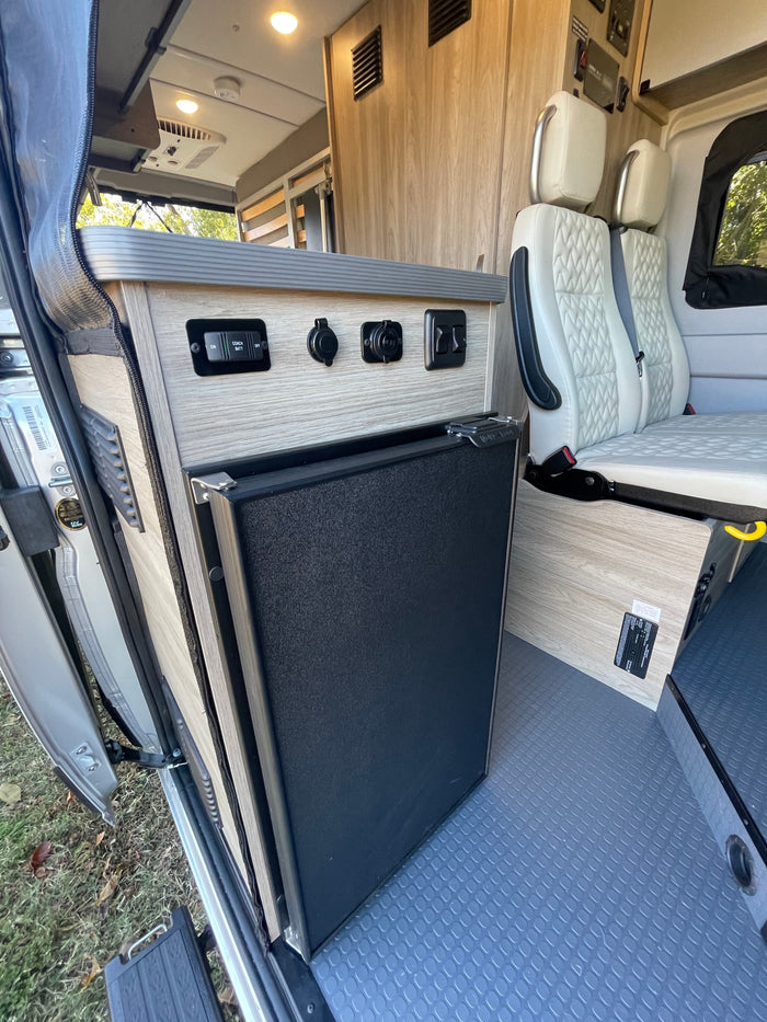 Inside an RV rental: a compact refrigerator, control panel, and comfortable passenger seating in a camper van.