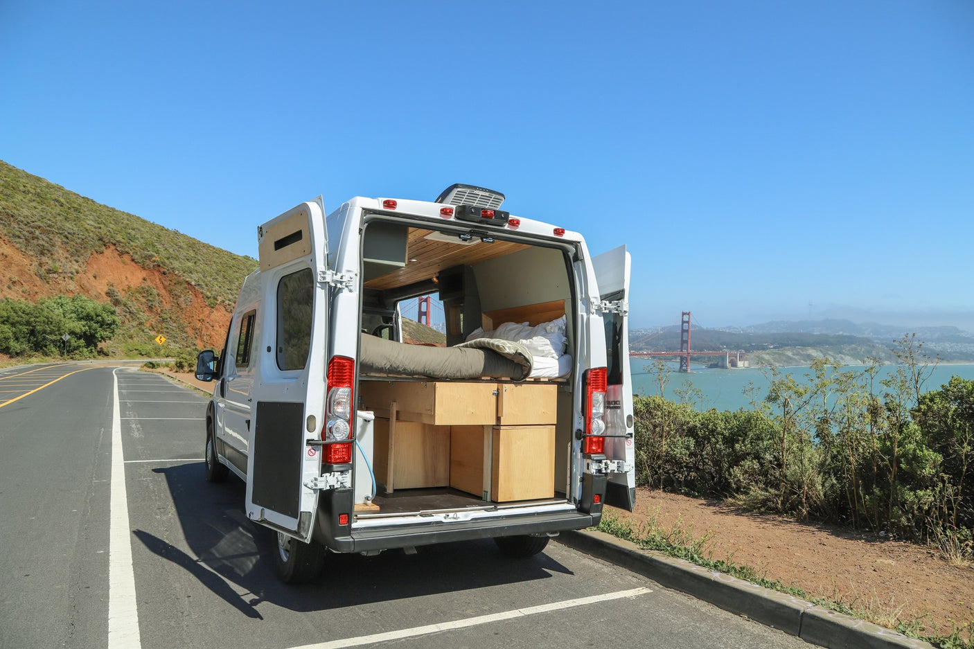 White RV rental camper van with open doors, revealing bed & storage, parked overlooking Golden Gate Bridge.