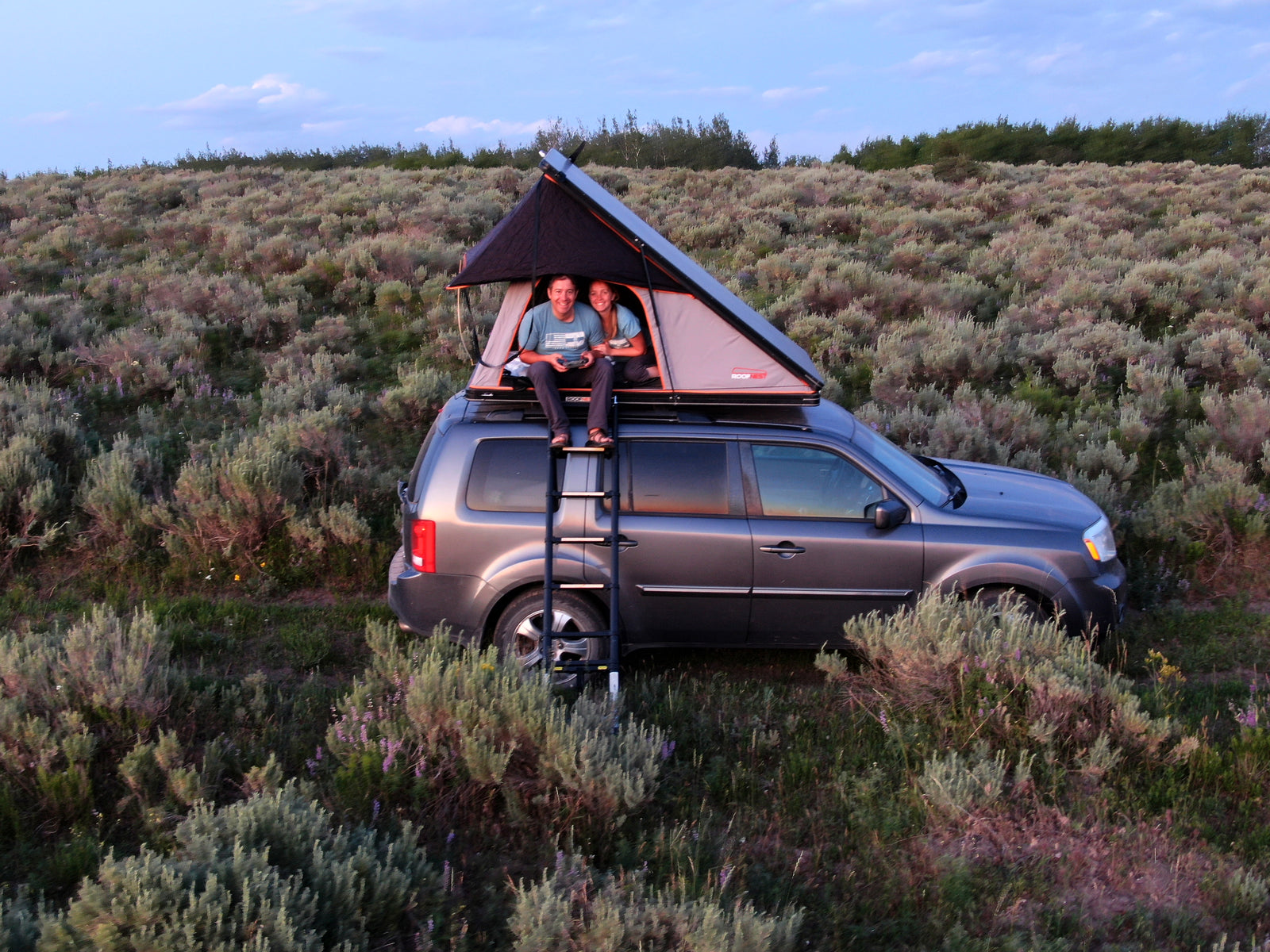Happy couple enjoys rooftop tent camping on their SUV, a compact alternative to an RV rental for a nature escape.