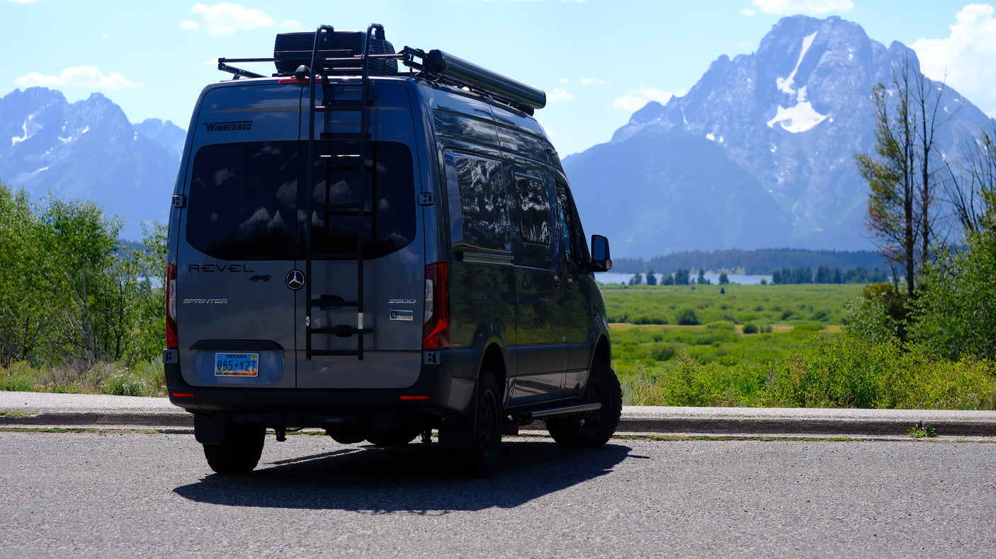 An RV rental: a gray Winnebago Revel camper van with roof ladder, backed onto asphalt, offering views of majestic mountains.