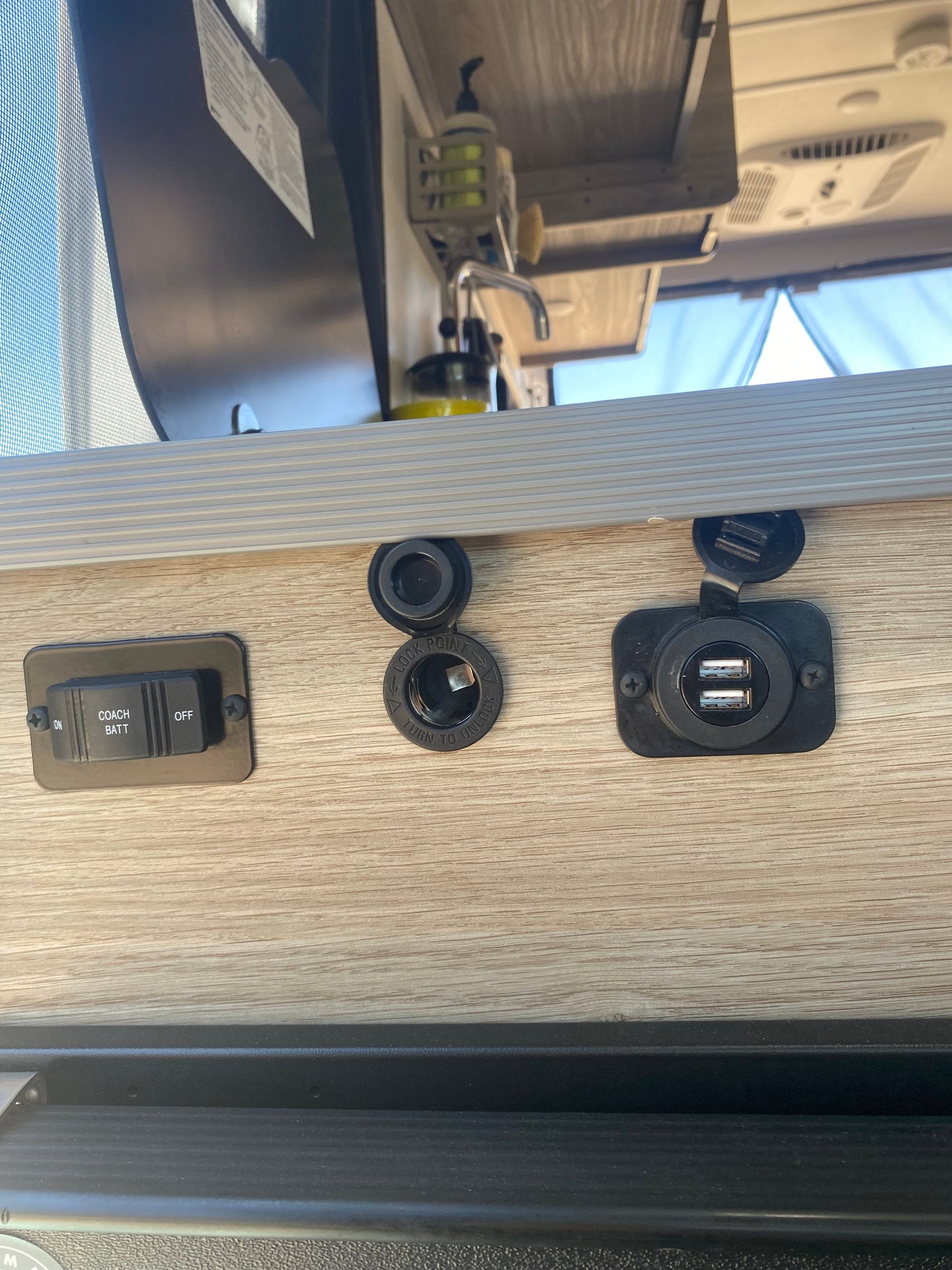 RV rental interior showing coach battery switch, 12V power point, and dual USB charging ports. Sink area above.