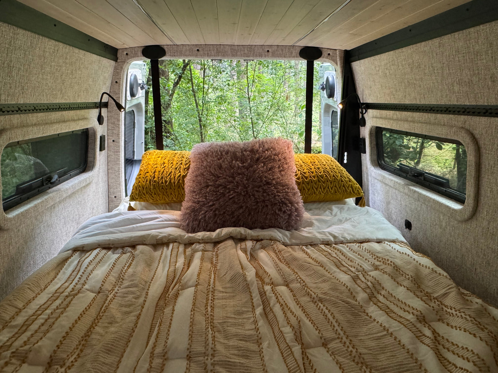 Step into comfort with this RV rental's bed, offering lush forest views from open back doors. Perfect for nature trips.