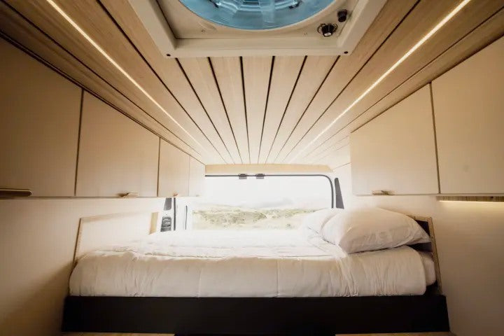 Experience van life! This modern RV rental interior features a cozy bed, wood ceiling with lighting, storage, and a scenic view.