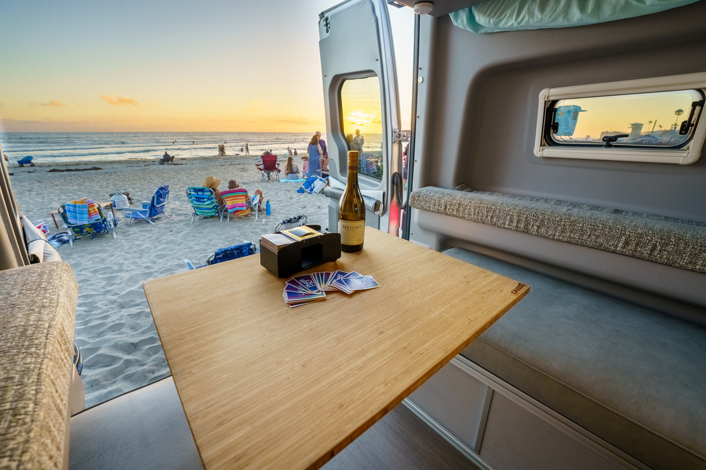 Enjoying a beach sunset from an RV rental. Inside a camper van, a bottle of wine & Phase 10 cards sit on a wooden table.
