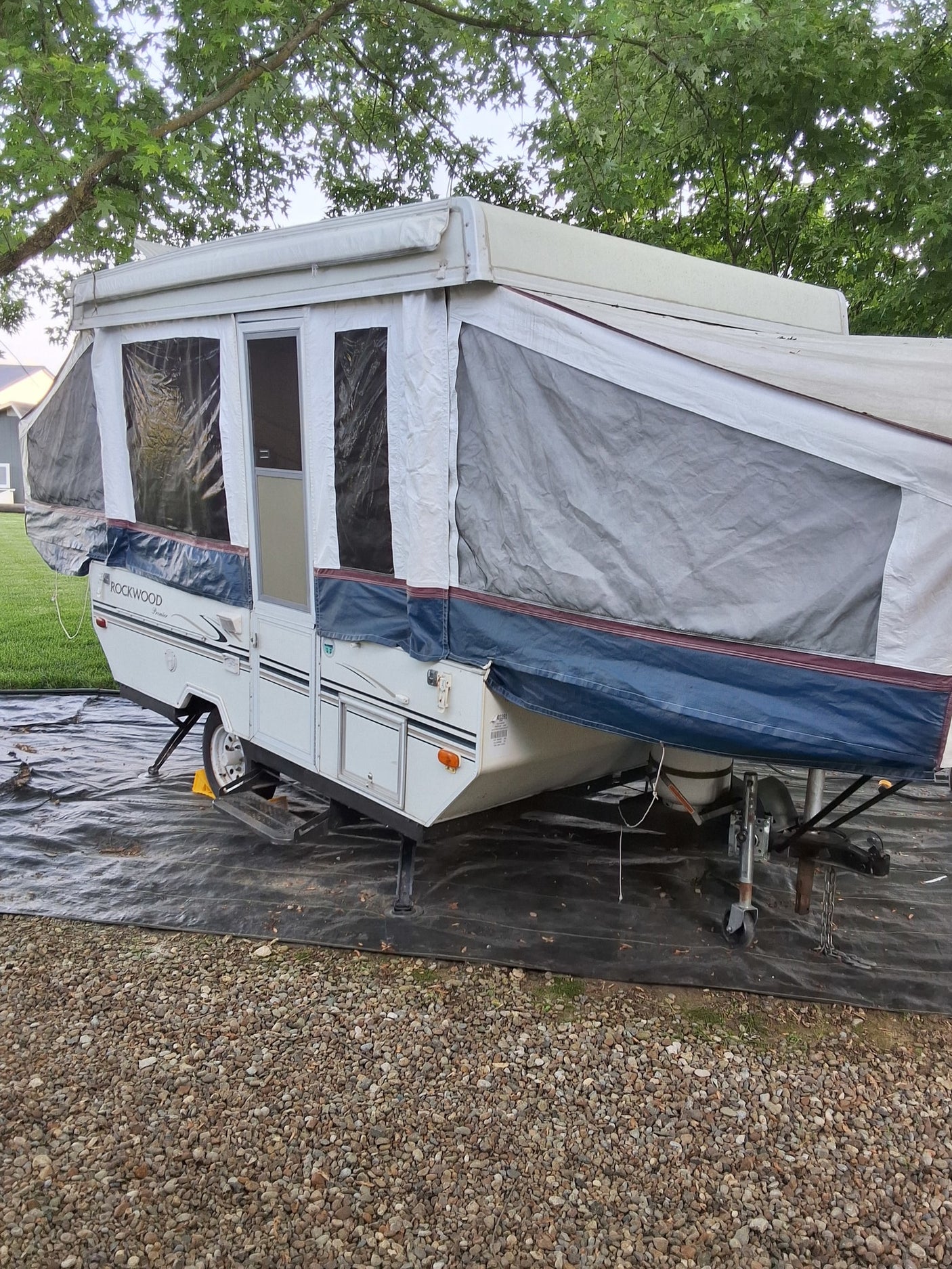 White Rockwood pop-up camper set up on gravel under trees. Ideal for an RV rental adventure.
