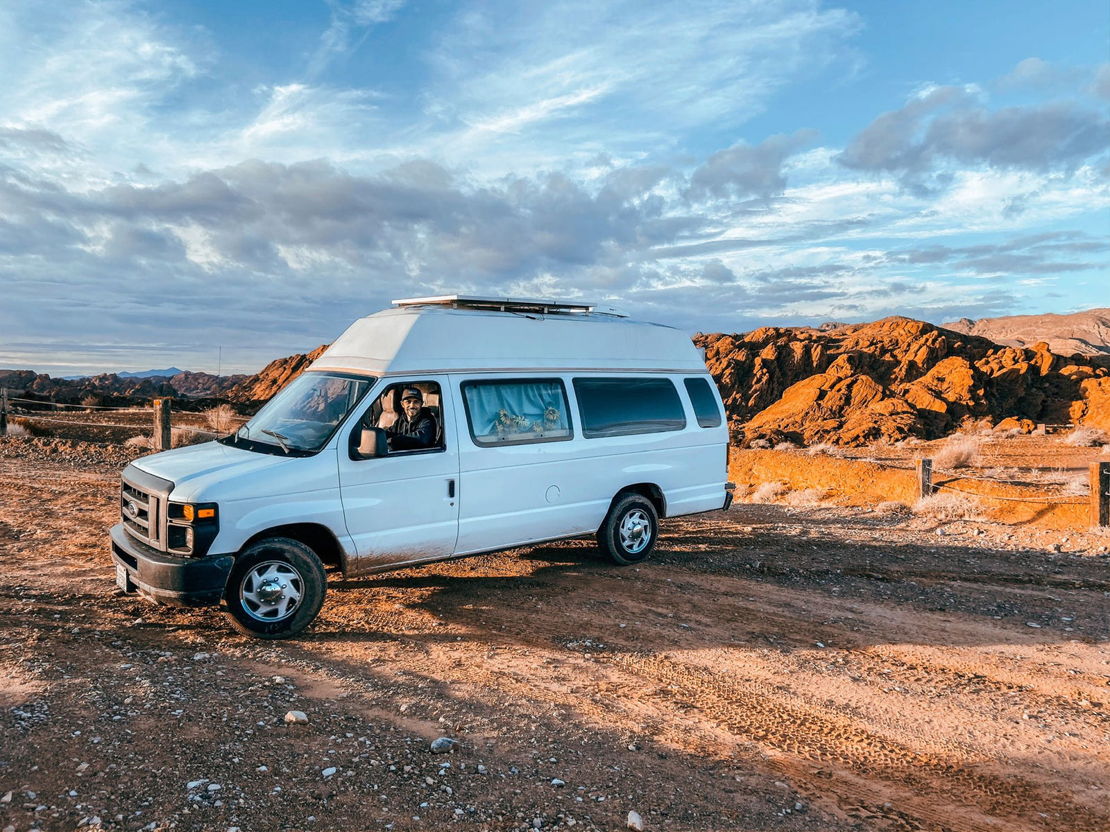 White van with high top, like an RV rental, parked on a dirt road in arid mountains under a dramatic sky. Person inside.