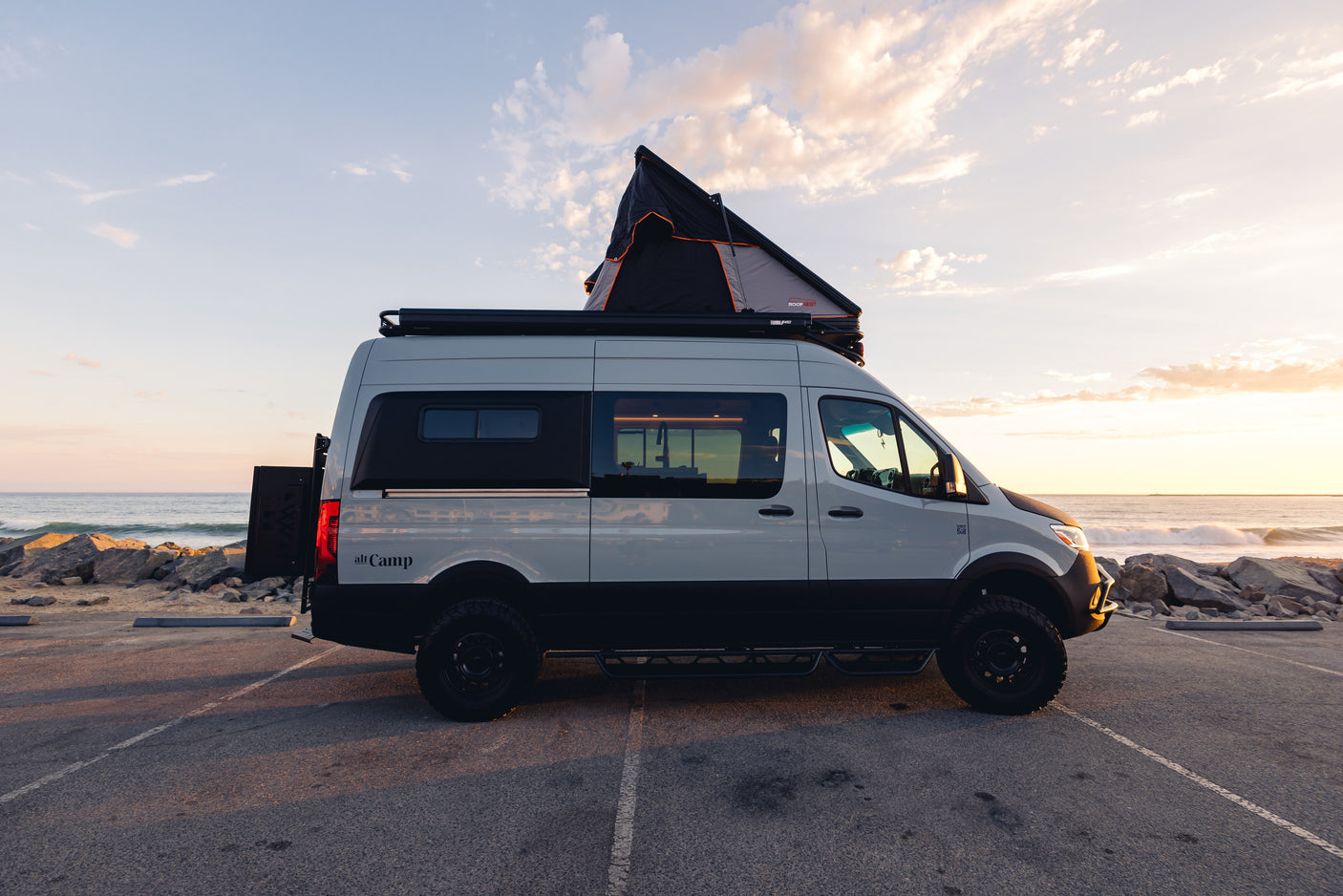 Custom two-tone RV rental camper van with rooftop tent, parked oceanfront at sunset. Perfect for adventure trips!