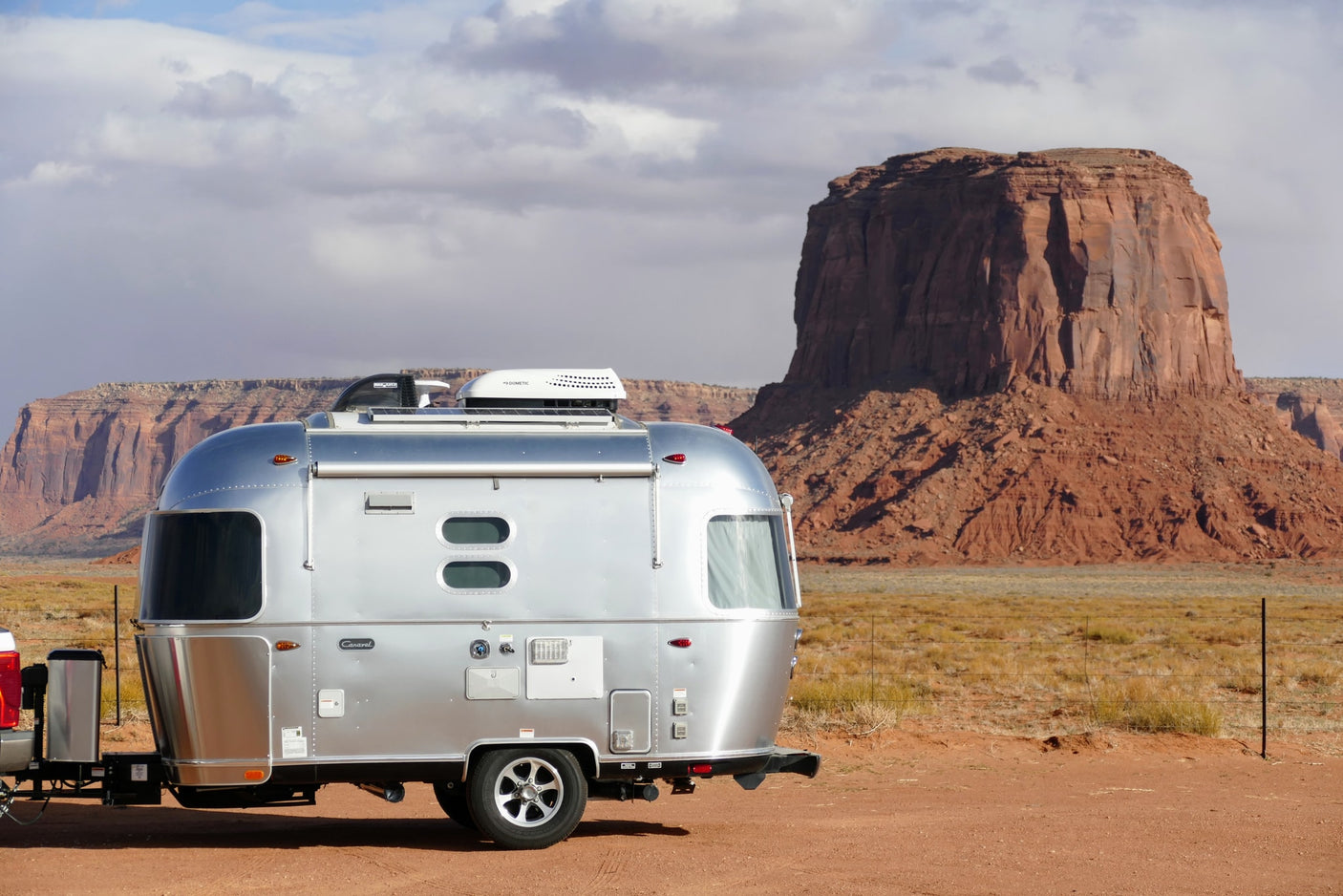 Silver Airstream travel trailer (RV rental) parked on red dirt, framed by towering red rock mesas under cloudy skies.