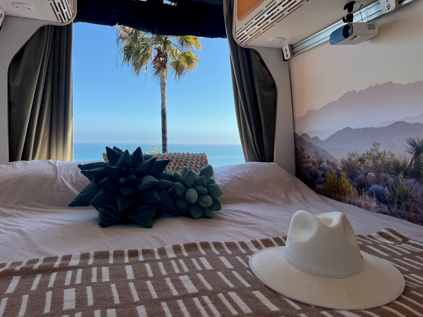 Cozy RV rental bed with ocean & palm tree views. Features succulent pillows, comfy blanket, and a ceiling-mounted projector for movies.