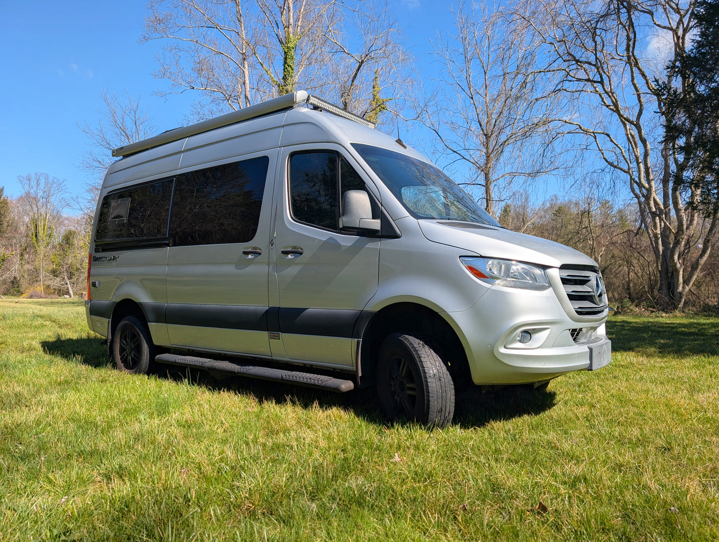 Silver Mercedes Sprinter RV rental parked on vibrant green grass under a clear blue sky, ideal for outdoor adventures.