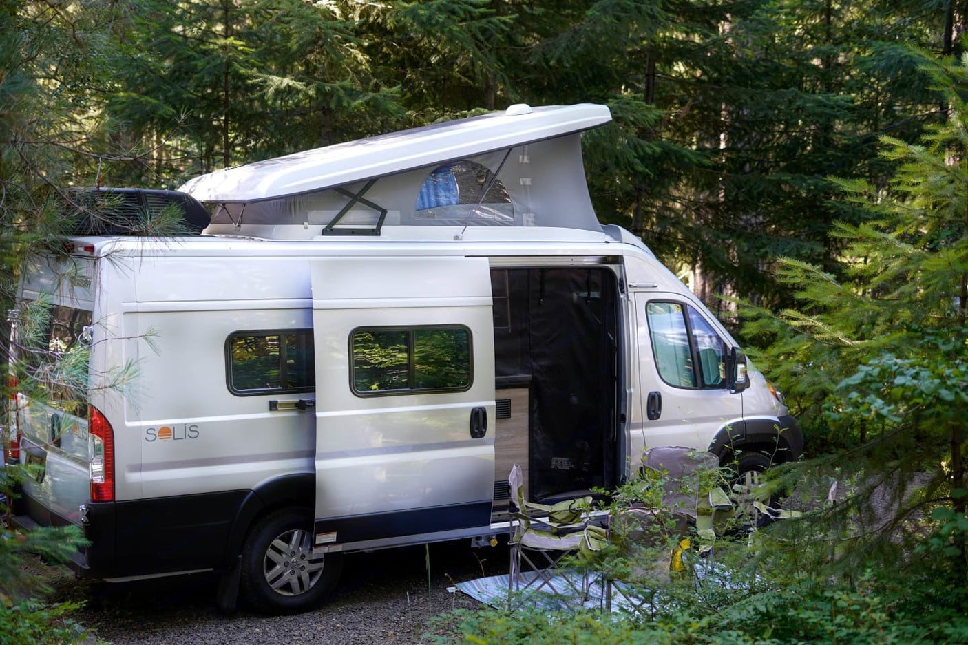 A bright Winnebago Solis rv rental van with an open pop-top and side door is parked among green trees, ready for camping.