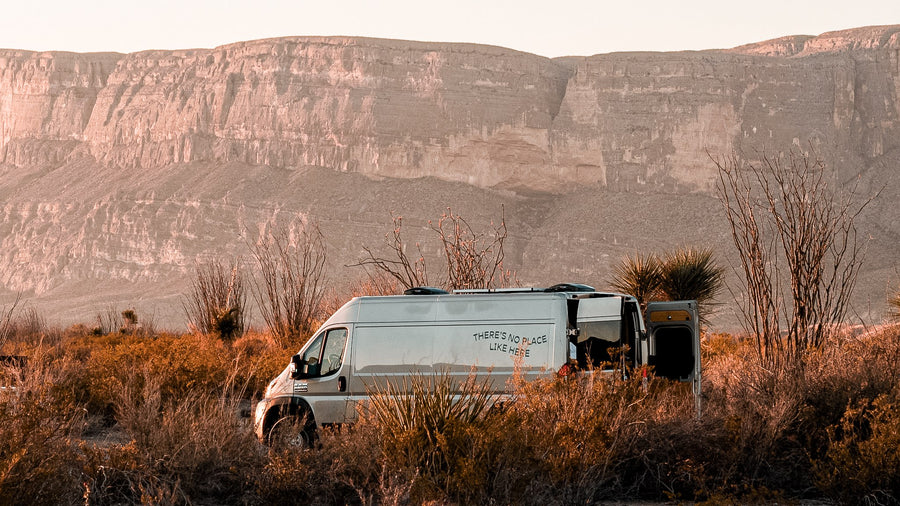 RV rental parked in a sunlit desert setting with towering mountains. The camper van's side reads 'NO PLACE LIKE HERE'.|Inside a stylish rv rental: light wood interior