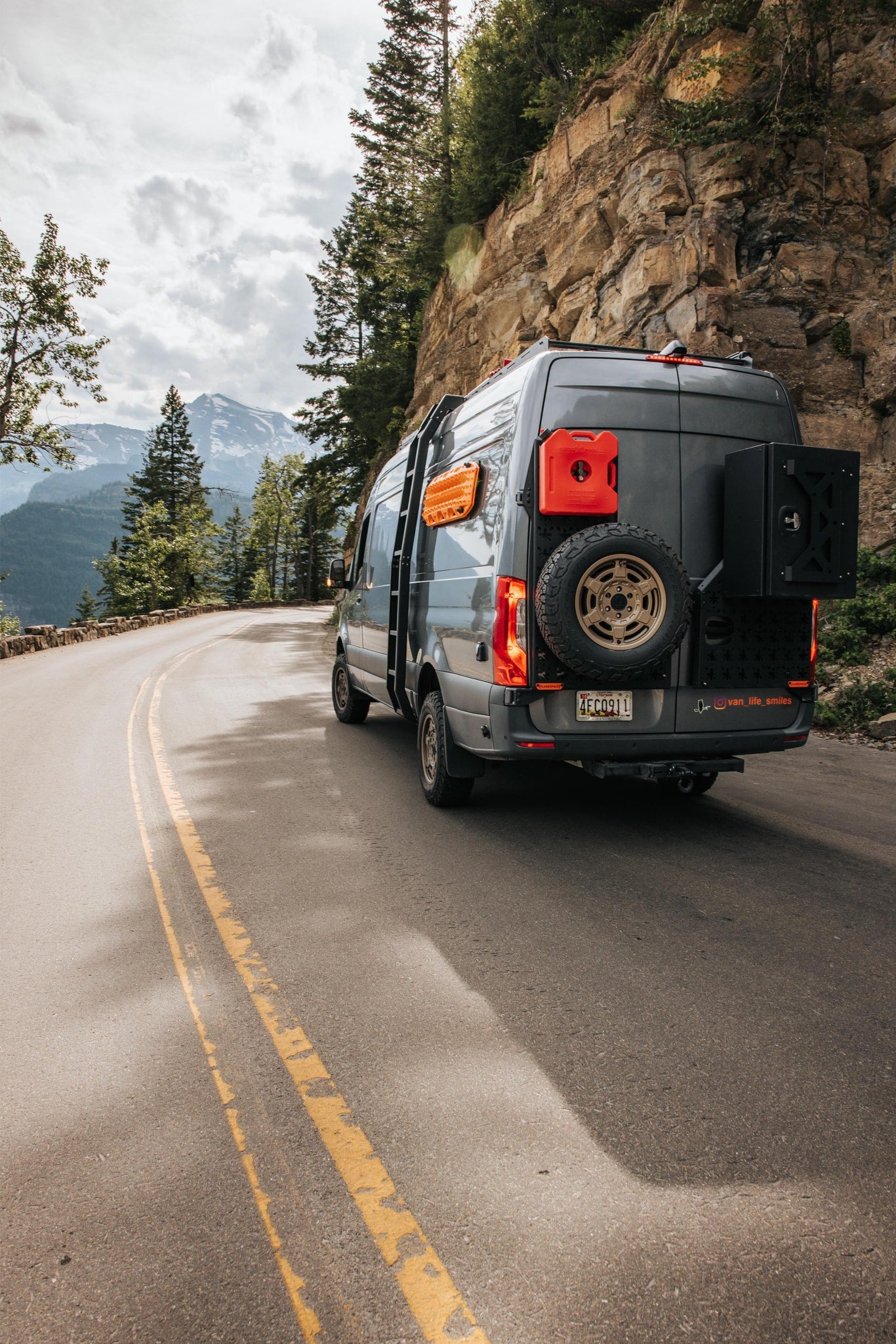 overlooking a stunning mountain panorama.|Gray off-road camper van on a scenic mountain road with rocky cliffs and snow-capped mountains. Perfect for your RV rental adventure.|Grey camper van with custom interior open in a forest setting