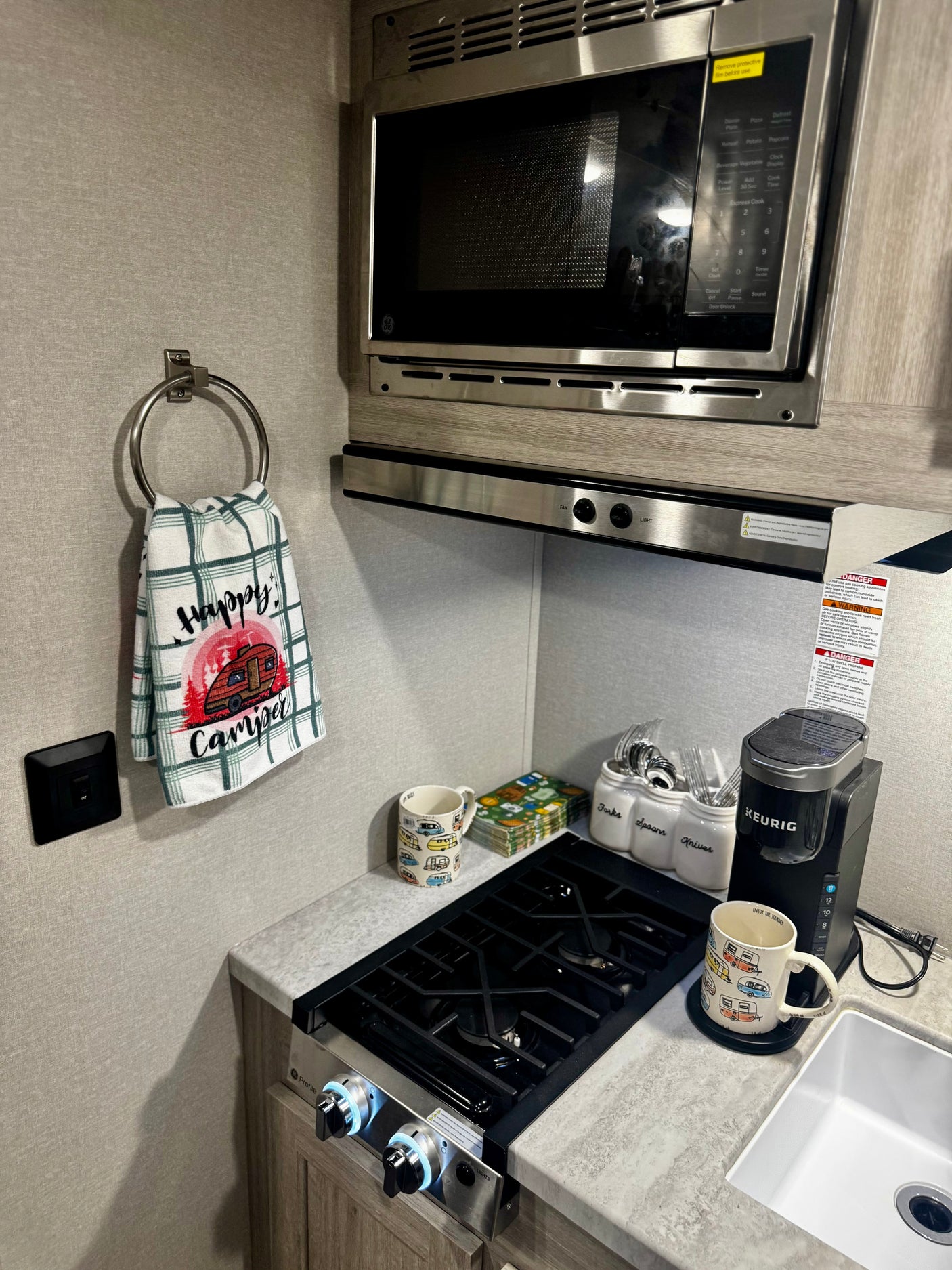 This RV rental kitchen features a microwave, stovetop, Keurig coffee maker, and "Happy Camper" decor, offering compact convenience.