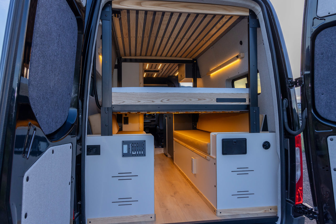 Inside an RV rental, a custom camper van features adjustable bunk beds, wood accents, and integrated controls, viewed from the rear.