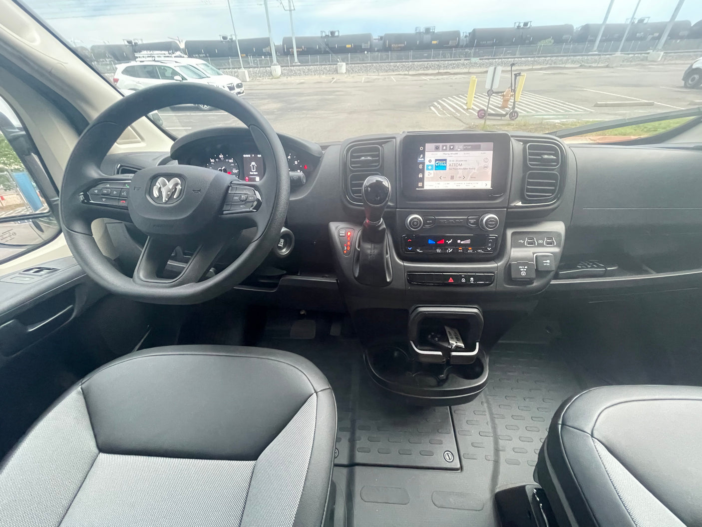 Inside a Ram ProMaster RV rental van: dashboard controls, steering wheel, and a clear view of a parking lot and train cars.