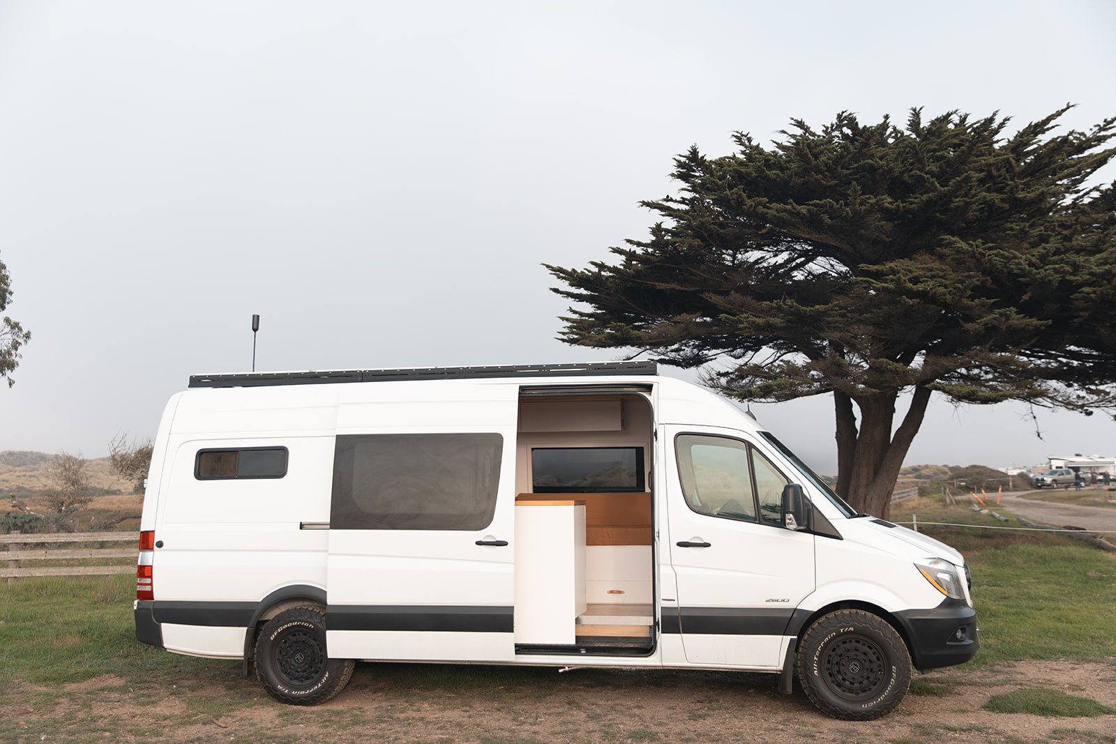 White Sprinter camper van for rv rental, ready for travel, open door reveals custom interior by a tree.