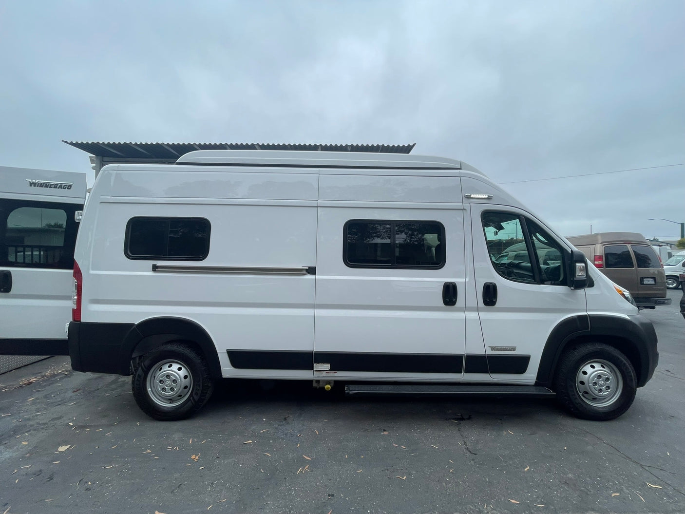 A white Winnebago camper van, shown in a side profile view, parked outdoors on an overcast day. Available for rv rental adventures.