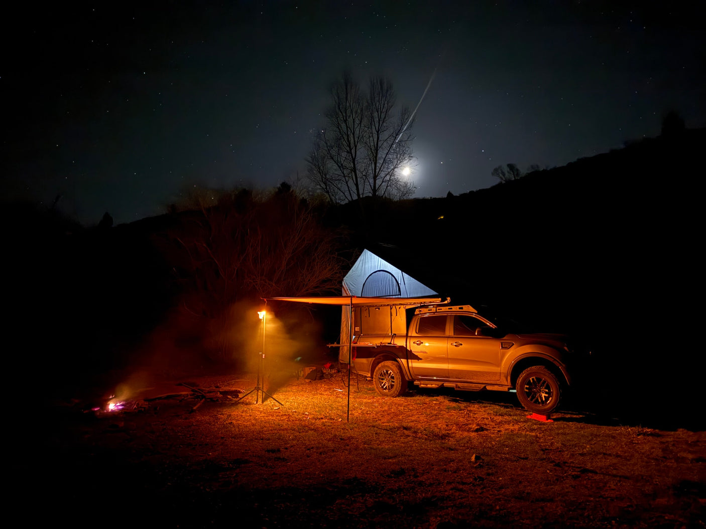 Starry night camping scene: a truck with a rooftop tent, awning, and campfire, under a bright moon. An ideal RV rental adventure.