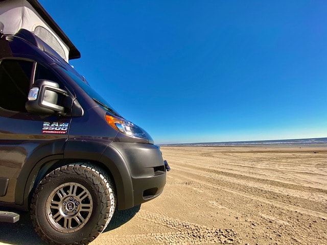 enjoying a breathtaking beach sunset. Ideal for travel & relaxation.|Experience beach adventures with this **RV rental**! A dark grey camper van with a pop-top roof is parked on a sandy beach at sunset.|This RV rental adventure features a dark gray van with pop-top