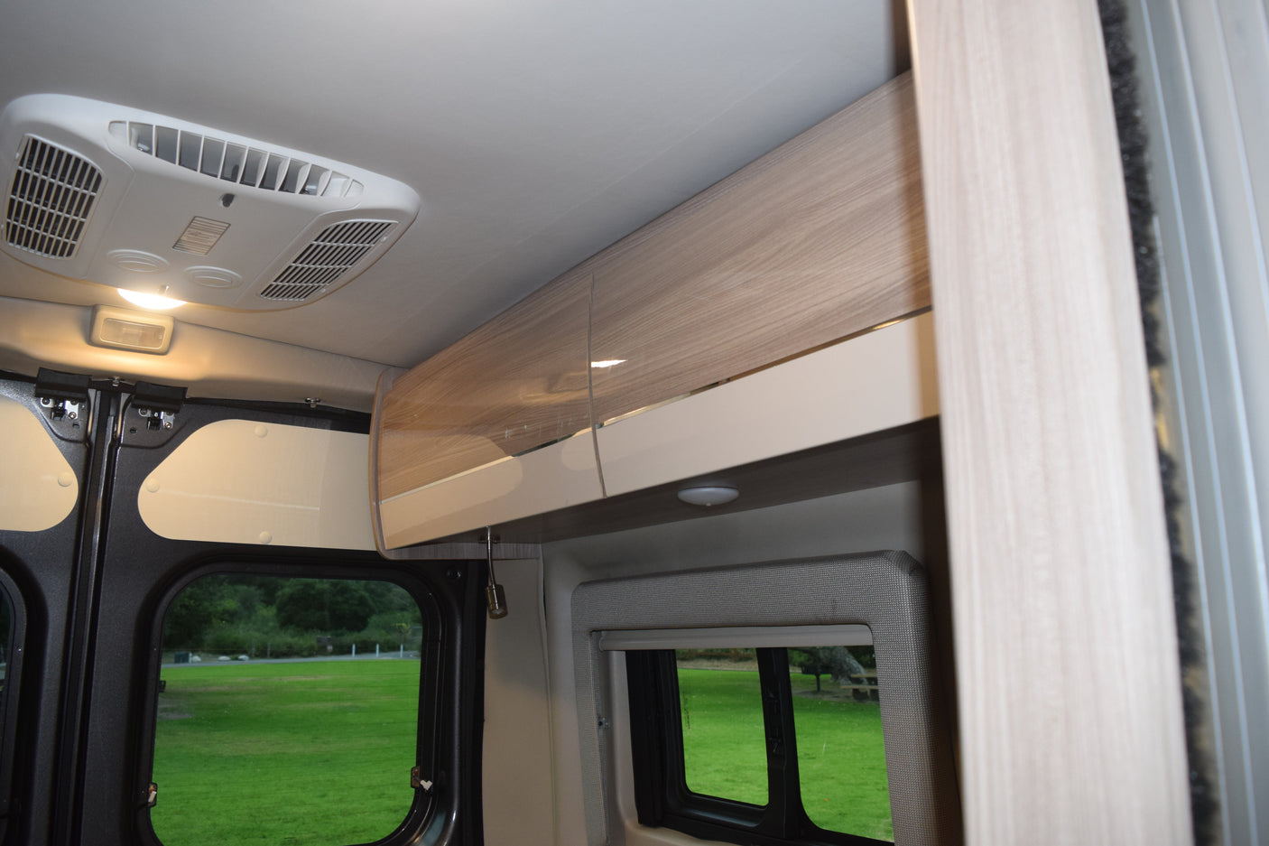 RV rental interior featuring an overhead AC unit, light wood-grain cabinets, and large windows overlooking a green campsite.